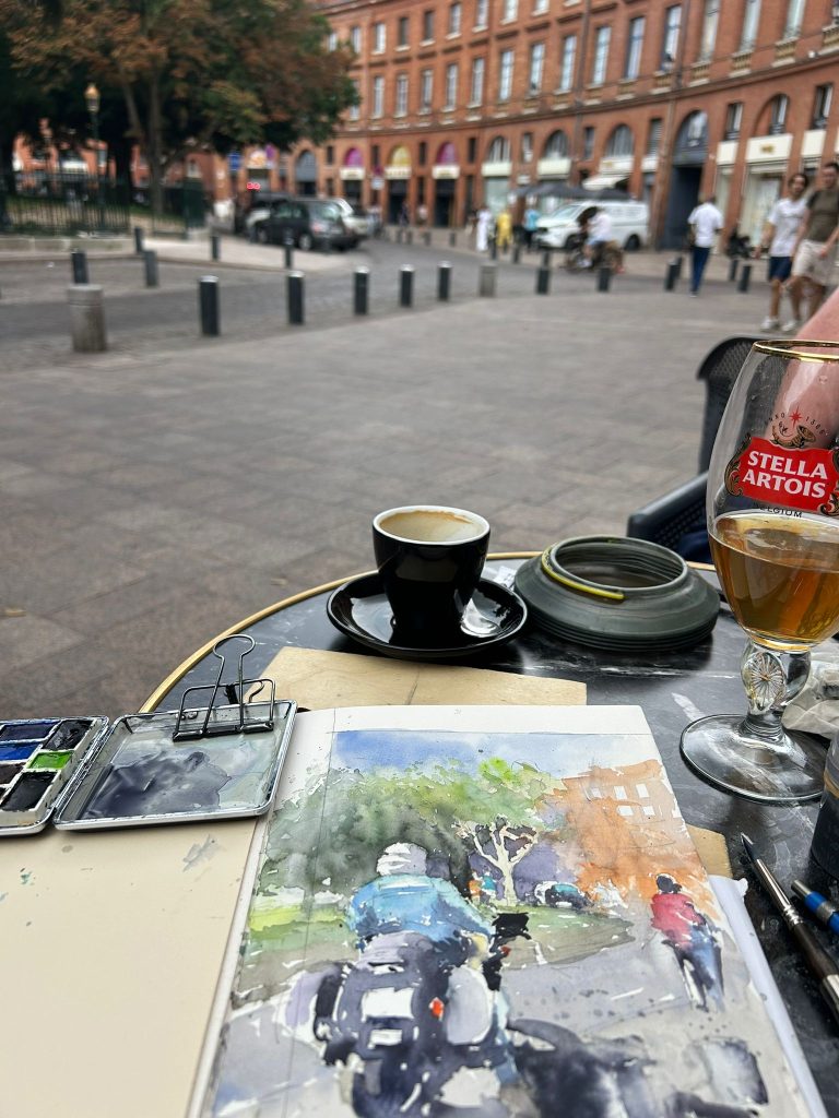 Cuaderno de acuarela y paleta sobre una mesa de terraza frente a una plaza